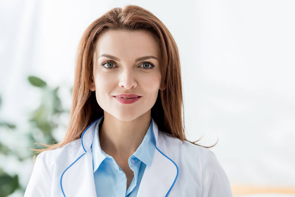 glimlachende arts in witte jas kijkend naar camera in ziekenhuis  - Foto, afbeelding