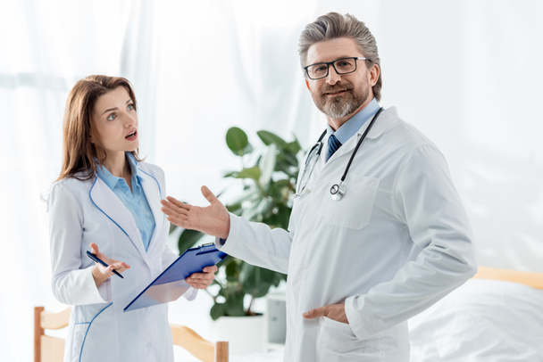 χαμογελαστός γιατρός με λευκό παλτό δείχνει με το χέρι και σοκαρισμένος συνάδελφος κοιτάζοντας τον στο νοσοκομείο - Φωτογραφία, εικόνα