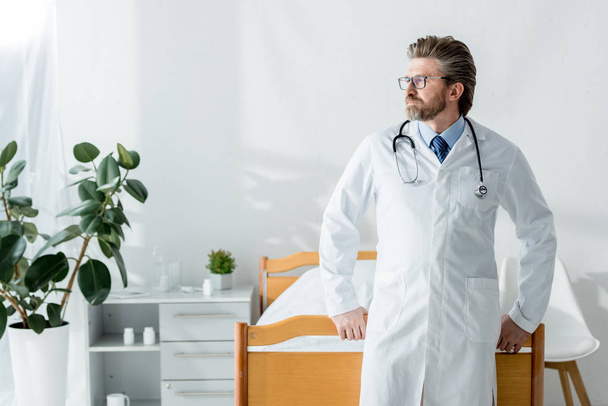 handsome doctor in white coat looking away in hospital  - Фото, изображение