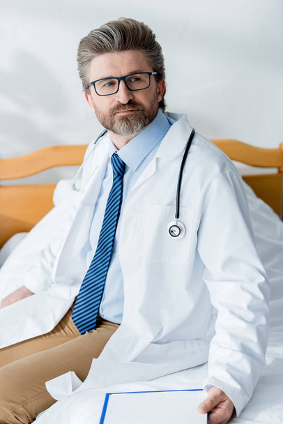 handsome doctor in white coat sitting on bed and looking away in hospital  - Fotografie, Obrázek
