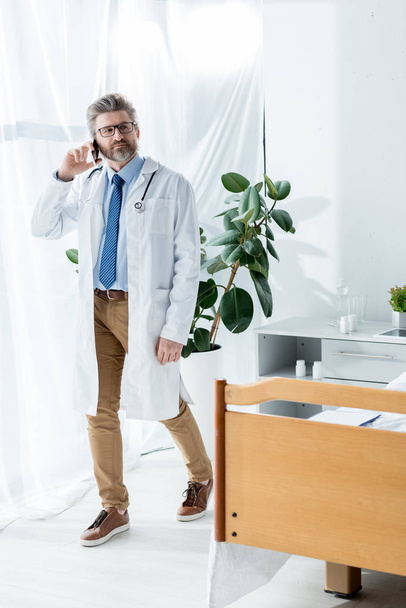 przystojny lekarz w białym płaszczu rozmawiający na smartfonie w szpitalu  - Zdjęcie, obraz