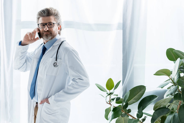 handsome doctor in white coat with hand in pocket talking on smartphone in hospital  - Photo, Image