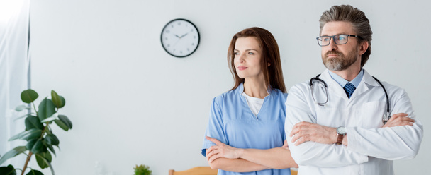 panoramisch schot van arts in witte jas en aantrekkelijke verpleegster met gekruiste armen weg te kijken in het ziekenhuis  - Foto, afbeelding