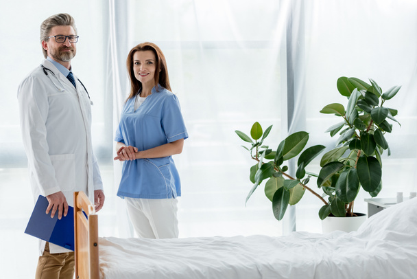 mosolygós orvos fehér kabátban és vonzó nővér nézi kamera a kórházban  - Fotó, kép