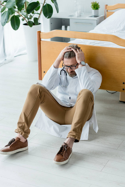 triste médico de bata blanca sentado en el suelo y tocando la cabeza en el hospital
  - Foto, imagen