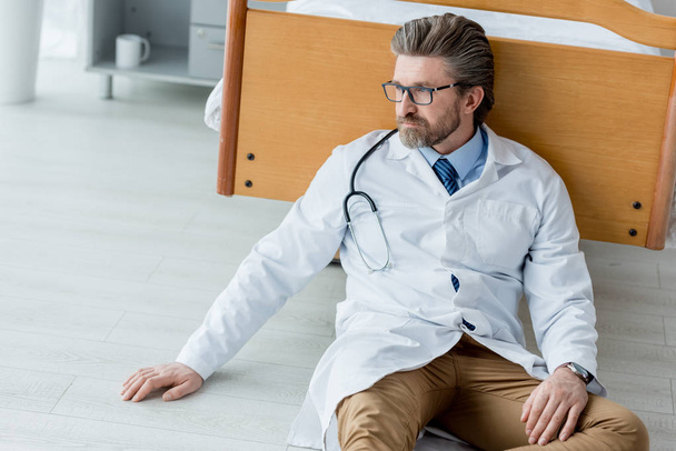 handsome doctor in white coat sitting on floor and looking away in hospital  - Photo, Image
