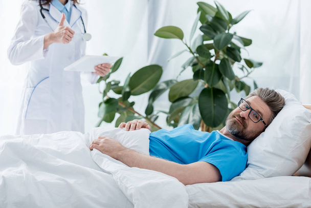 cropped view of doctor in white coat showing like and holding digital tablet and patient lying in bed in hospital  - Photo, Image