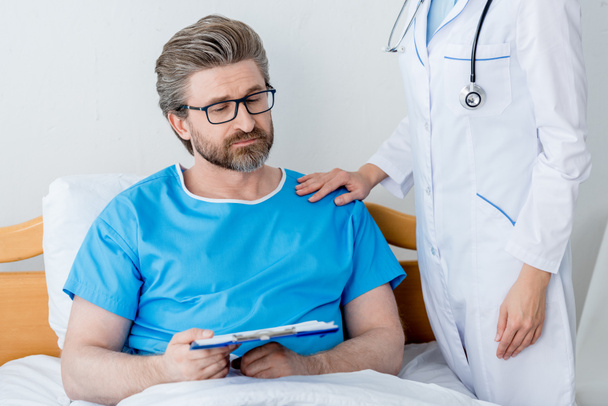 vista recortada del médico de bata blanca tocando hombro de paciente triste con portapapeles
  - Foto, Imagen