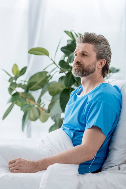 handsome patient in medical gown looking away in hospital  - Fotografie, Obrázek