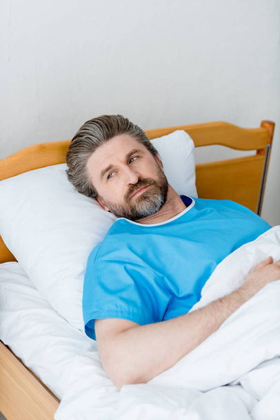 θέα υψηλής γωνίας του ασθενούς με ιατρική ρόμπα ξαπλωμένος και κοιτάζοντας μακριά στο νοσοκομείο  - Φωτογραφία, εικόνα