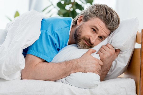 guapo paciente en bata médica acostado en la cama y mirando hacia otro lado
  - Foto, Imagen