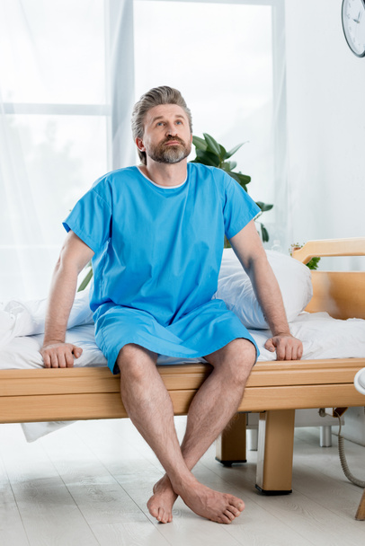 ασθενής με ιατρική ρόμπα κάθεται στο κρεβάτι και ψάχνει στο νοσοκομείο  - Φωτογραφία, εικόνα