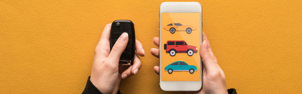 oříznutý pohled na ženu držící klíč od auta a smartphone s půjčovnou aut app na obrazovce na oranžovém pozadí, panoramatický záběr - Fotografie, Obrázek