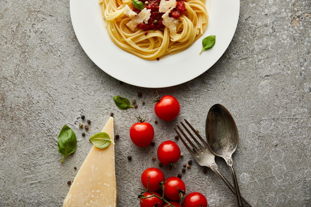 Draufsicht auf köstliche Spaghetti mit Tomatensauce auf Teller in der Nähe von Käse, Tomaten und Besteck auf grau strukturierter Oberfläche - Foto, Bild