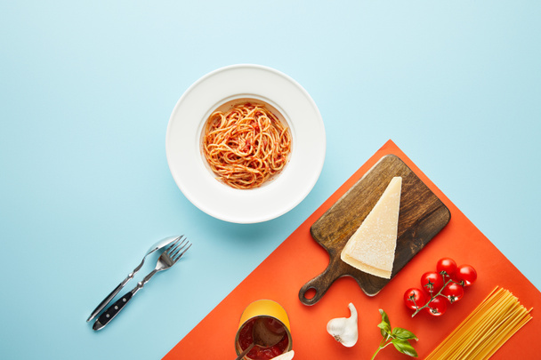 flat lay with delicious spaghetti with tomato sauce in plate near cutlery and ingredients on blue, red background - Photo, Image