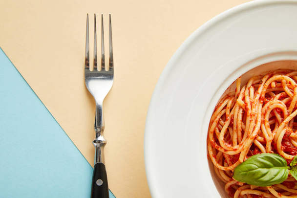 top view of delicious spaghetti with tomato sauce in plate near fork on blue and yellow background - Foto, afbeelding