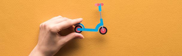 cropped view of woman holding paper cut kick scooter on orange background, panoramic shot - 写真・画像