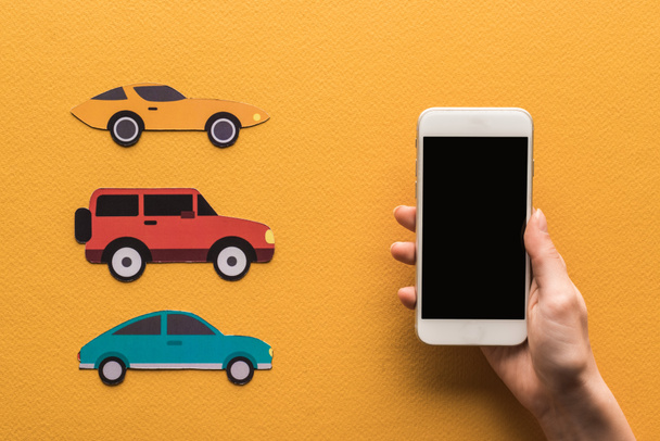 bijgesneden weergave van vrouw met smartphone met blanco scherm in de buurt van papier gesneden auto 's, locatie merk op oranje achtergrond - Foto, afbeelding