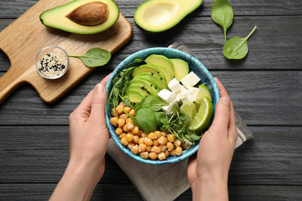Woman holding bowl of delicious avocado salad with chickpea at black wooden table, top view - Foto, Bild