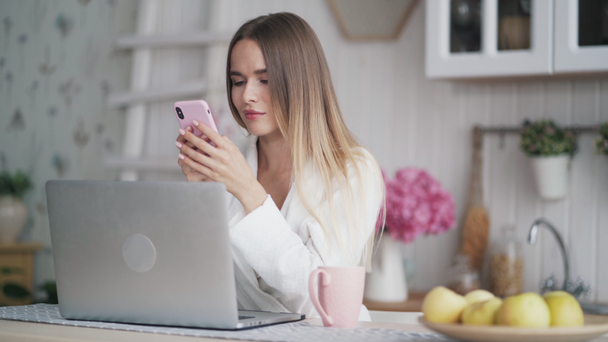 Mujer joven se sienta en la mesa con el ordenador portátil en la cocina, utiliza el teléfono para chatear en línea
 - Imágenes, Vídeo