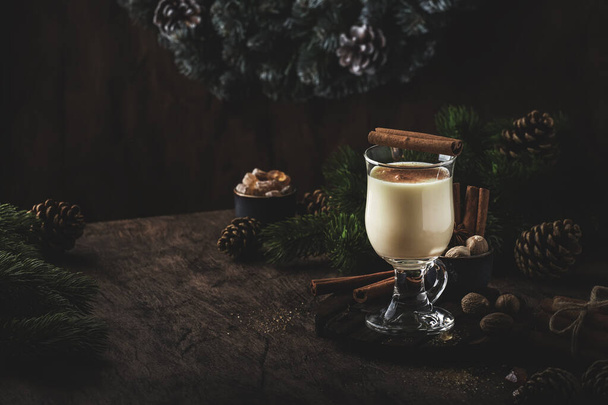 New Year or Christmas Eggnog cocktail - hot winter or autumn drink with milk, eggs and dark rum, sprinkled with cinnamon and nutmeg in a glass on wooden background, festive decoration - Foto, Imagem