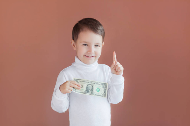 malý chlapec drží v rukou jeden dolar na růžovém pozadí, radost z prvních výdělků, dobrý příspěvek pro dítě do budoucna - Fotografie, Obrázek