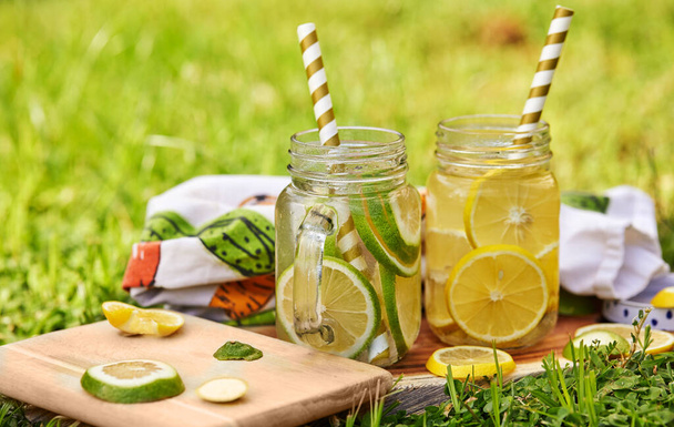 Δροσιστικό ποτό με λεμόνι και λάιμ σε ειδικά βάζα με καλαμάκια σε ξύλινο σταντ, τραβηγμένο το πρωί στο πράσινο γρασίδι. Έννοια υγιεινής διατροφής - Φωτογραφία, εικόνα
