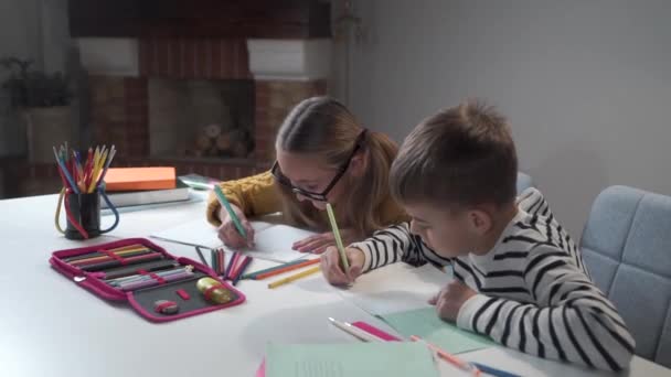 Caucasian brother and sister doing homework together. Joyful children writing in siblings exercise book. Boy in striped jacket and girl in eyeglasses sitting at the table at home and doing tasks. - Кадры, видео