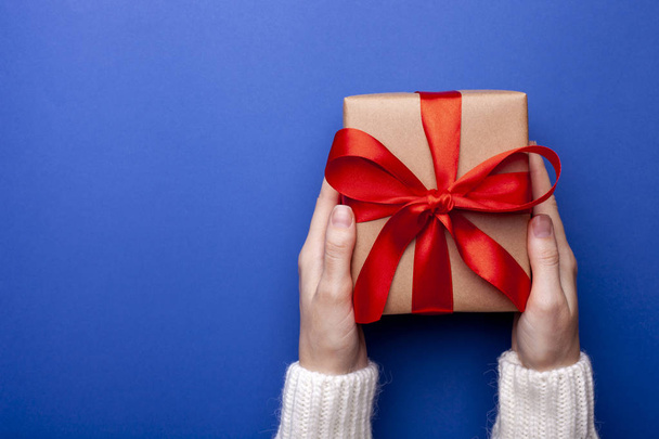 Egy nő ajándékot tart karácsonyra a kék háttérben. Női kéz kezében ajándék doboz csomagolva kézműves papír piros szalaggal színes háttér. Felülnézet szöveges hellyel - Fotó, kép