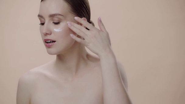 attraktive Frau, die Gesichtscreme isoliert auf Beige aufträgt  - Filmmaterial, Video