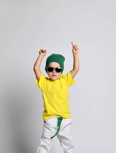 fröhlicher flippiger kleiner Junge hält Hände über seinem Kopf frei Textkopierraum. gelbes T-Shirt und grüner Hut. - Foto, Bild