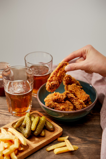 cropped view of woman eating delicious chicken nuggets, french fries and gherkins near glasses of beer on wooden table isolated on grey - Foto, Bild