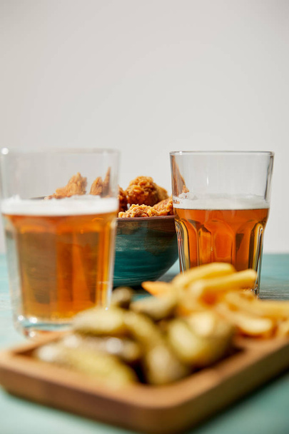foco seletivo de deliciosas pepitas de frango, batatas fritas e pepinos perto de copos de cerveja em mesa de madeira turquesa em fundo cinza
 - Foto, Imagem