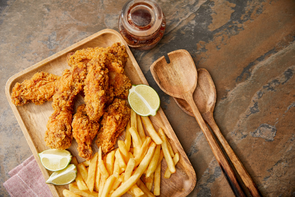 top view of wooden board with delicious chicken nuggets, french fries and lime on stone surface with spatulas - Foto, immagini