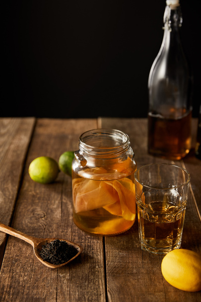 Kireç, limon, baharat ve şişe yanında kombu çayı olan cam kavanozun seçici odak noktası, siyah üzerine izole edilmiş ahşap masa. - Fotoğraf, Görsel