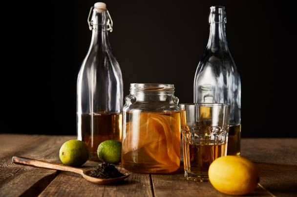 glass jar with kombucha near lime, lemon, spice and bottles on wooden table isolated on black - Foto, Imagem