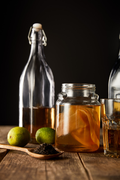 glass jar with kombucha near lime, spice and bottle on wooden table isolated on black - Photo, Image