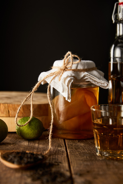selective focus of glass jar with kombucha near lime, spice and bottle on wooden table isolated on black - Zdjęcie, obraz