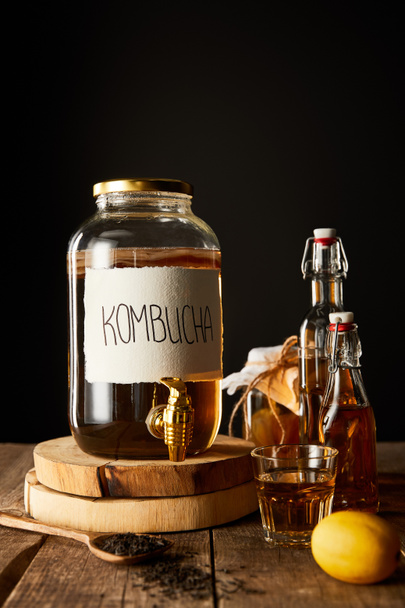 frasco de vidrio con kombucha cerca de limón, especias y botellas en mesa de madera aislado en negro
 - Foto, Imagen