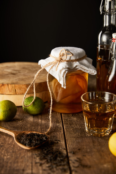 szklany słoik z kombucha w pobliżu limonki, cytryny, przyprawy i butelki na drewnianym stole odizolowany na czarno - Zdjęcie, obraz