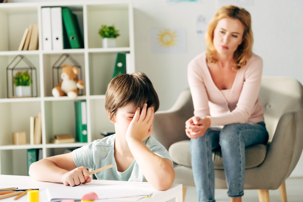 trauriges Kind mit Legasthenie mit Bleistift und Kinderpsychologe im Hintergrund  - Foto, Bild