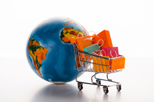 toy shopping cart with shopping bags near globe on white, e-commerce concept - Foto, afbeelding