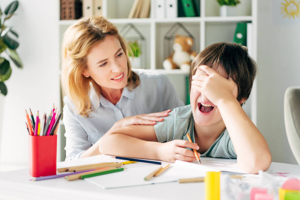 upset kid with dyslexia obscuring face and crying, smiling child psychologist looking at him  - Photo, Image