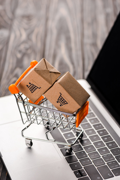 selective focus of toy carton boxes in small shopping cart on laptop with blank screen, e-commerce concept - Photo, Image
