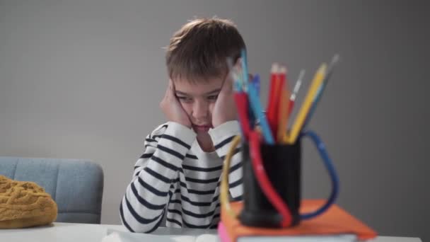 Exhausted Caucasian sleepy boy sitting at the table at school. Schoolboy holding his head with hands. Tired bored child sitting at lessons. Difficulties in education process. - Video