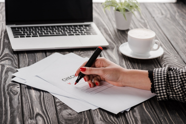 обрезанный вид деловой женщины, держащей маркер ручку возле контрольного списка, чашку и ноутбук на столе, концепция электронной коммерции
 - Фото, изображение
