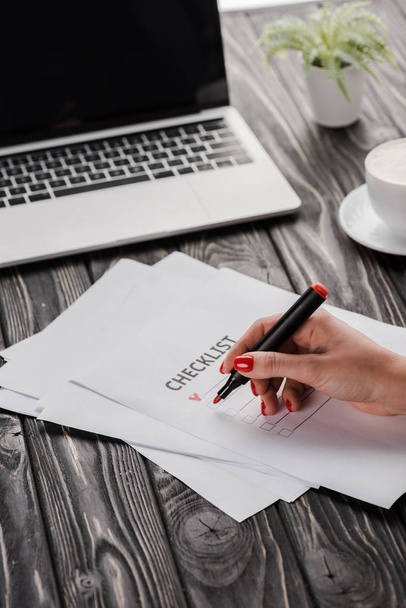 vágott kilátás üzletasszony kezében piros marker toll közelében ellenőrzőlista, csésze és laptop az asztalon, e-kereskedelmi koncepció - Fotó, kép