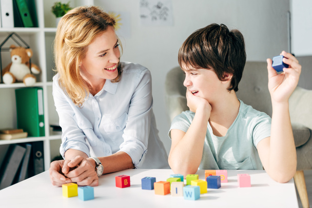 улыбающийся детский психолог смотрит на ребенка с дислексией и сидит за столом со строительными блоками
 - Фото, изображение