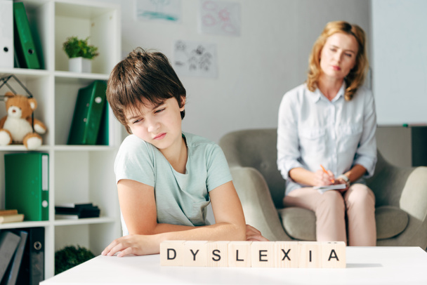 enfoque selectivo de niño triste con dislexia sentado en la mesa con cubos de madera con letras dislexia
 - Foto, Imagen