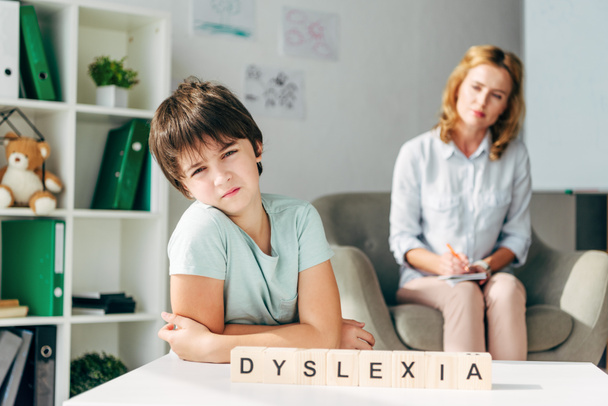 selective focus of sad kid with dyslexia sitting at table with wooden cubes with lettering dyslexia - Photo, Image